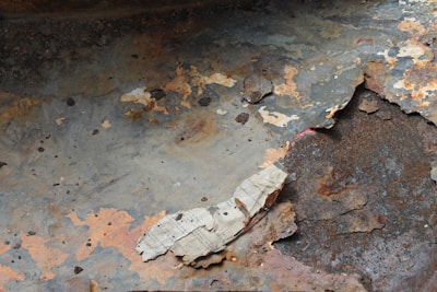 Peeling and rusted metal surface with layers of paint flaking off. The underlying rust and corrosion are visible, showing various shades of brown and orange. There are patches where the paint has completely peeled away, exposing the raw metal. The texture is rough and uneven.