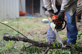 A person wearing gloves is operating an orange chainsaw to cut through a fallen tree branch. The surroundings include grass and some out-of-focus background elements, such as a structure and various tools.
