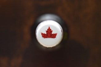 A close-up view of the cap of a bottle, featuring a red maple leaf design on a white background. The cap is centered and the overall scene has a soft focus with a dark, blurred background.