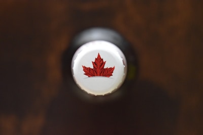 A close-up view of the cap of a bottle, featuring a red maple leaf design on a white background. The cap is centered and the overall scene has a soft focus with a dark, blurred background.