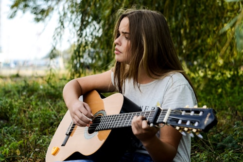 A young woman with long brown hair sits outdoors among green foliage, playing an acoustic guitar. She is wearing a light-colored t-shirt, and her expression appears thoughtful or focused.