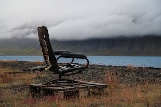 A solitary, worn-out chair sits atop a wooden pallet on a grassy, rocky shore with a misty lake and a mountain range in the background. The clouds hang low, enveloping the mountains partially in a soft, gray blanket.