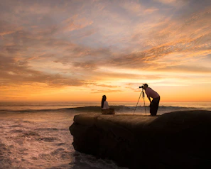 A candid portrait of Radit adjusting his camera on a rocky coastline during sunset