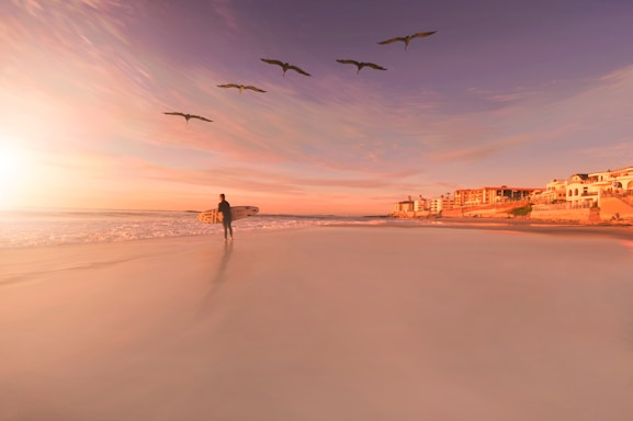 A serene beach at sunset with a lone surfer carrying a board along the shore.