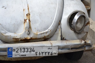 The image depicts the front part of a vintage car, characterized by a rusty, worn-out appearance. The vehicle's paint is faded, and there are visible rust spots around the headlights and bumper. The license plate, marked 'LIBAN', indicates it's from Lebanon.