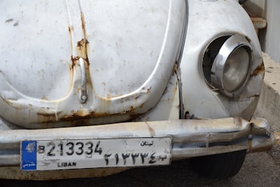 The image depicts the front part of a vintage car, characterized by a rusty, worn-out appearance. The vehicle's paint is faded, and there are visible rust spots around the headlights and bumper. The license plate, marked 'LIBAN', indicates it's from Lebanon.
