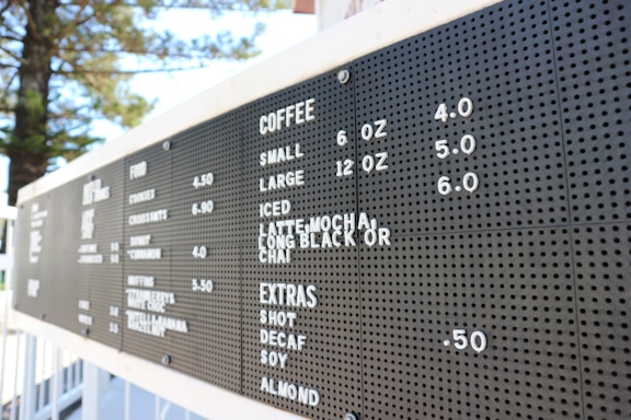 A black menu board listing coffee options with sizes and prices, as well as food and extras. The board is outdoors with trees visible in the background.