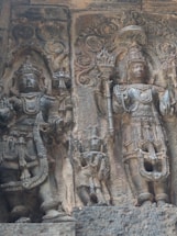 The image depicts an intricate stone carving featuring two deities adorned with elaborate headdresses and jewelry, set against a textured stone background. The deities are depicted standing, with one holding a trident-like object. Surrounding them is ornate detailing, showcasing traditional motifs and decorative elements.