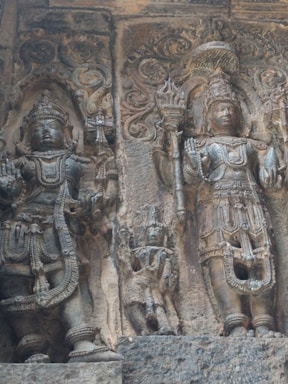 The image depicts an intricate stone carving featuring two deities adorned with elaborate headdresses and jewelry, set against a textured stone background. The deities are depicted standing, with one holding a trident-like object. Surrounding them is ornate detailing, showcasing traditional motifs and decorative elements.
