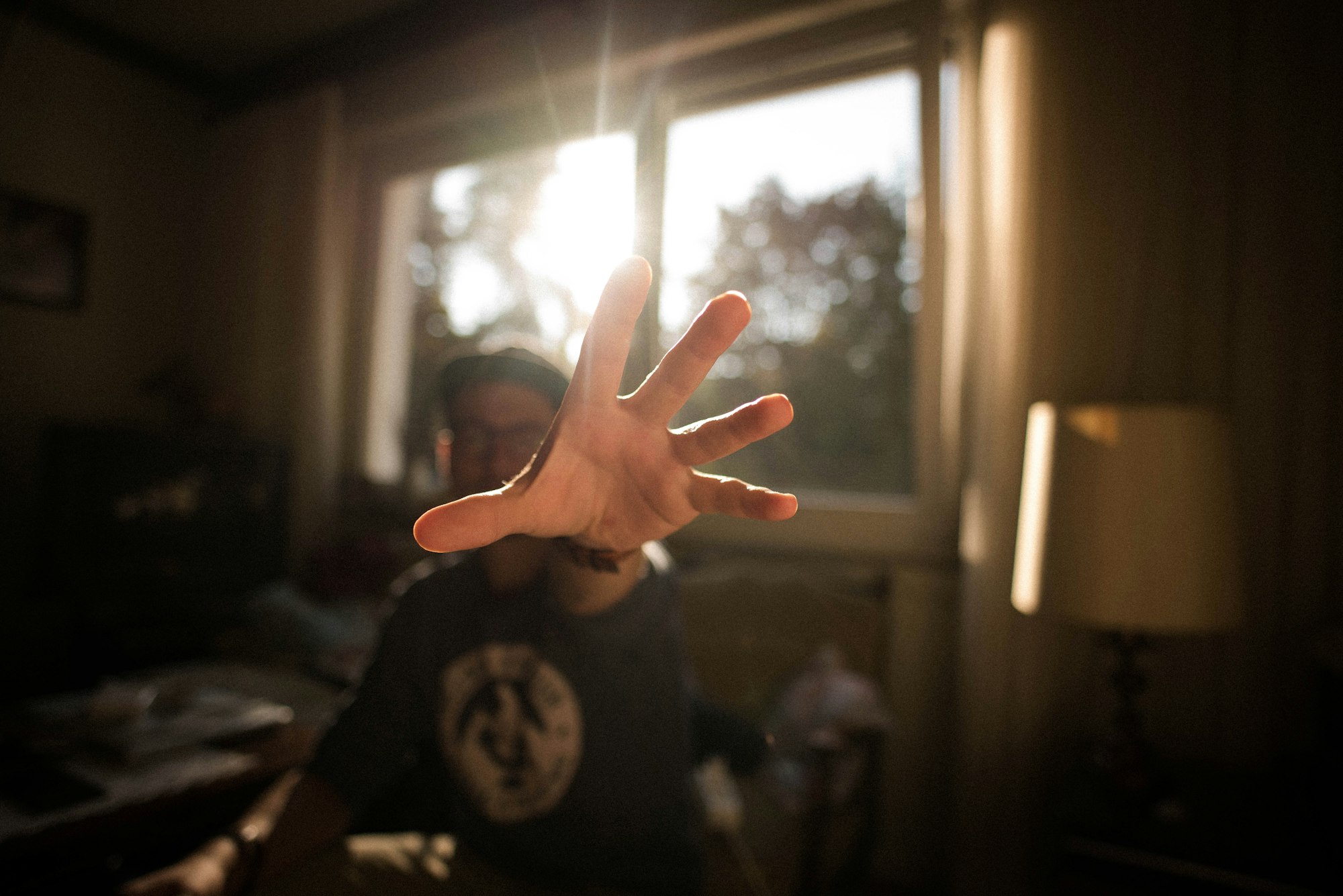 A man in a residroom holds an outstretched hand in front of the camera, with the sun appearing in a window in the background.