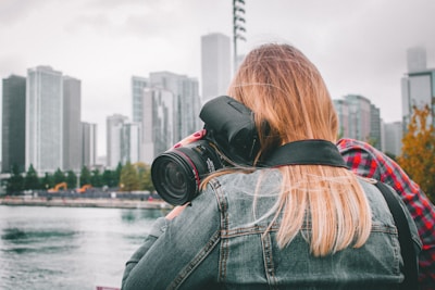 A person with long blonde hair is focused on taking a photograph using a large camera with a telephoto lens. The setting appears to be an urban environment with high-rise buildings in the background, a body of water, and some autumn trees. The individual is wearing a denim jacket, and there are overcast skies, adding a muted tone to the scene.