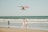 A bright red seaplane flies low over the ocean near the shore, with gentle waves rolling onto the beach. Several people are seen enjoying the water, including a couple standing at the water's edge and a child playing in the surf.