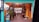 An artistic space with bright colors and decorative lighting. The entrance is framed with a teal and orange color scheme, and the area is adorned with potted plants and various artistic signs. A colorful trash can and a welcoming sign are visible, suggesting a cozy and creative environment.
