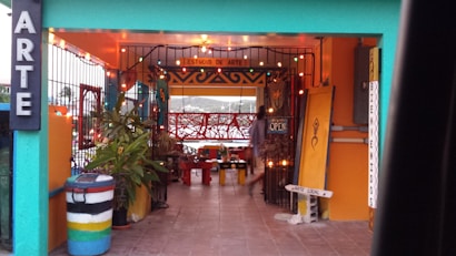 An artistic space with bright colors and decorative lighting. The entrance is framed with a teal and orange color scheme, and the area is adorned with potted plants and various artistic signs. A colorful trash can and a welcoming sign are visible, suggesting a cozy and creative environment.