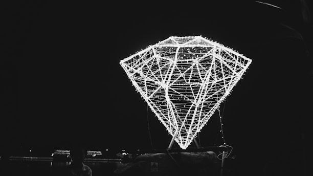 A large, illuminated structure in the shape of a diamond is constructed with bright lights against a dark background. The structure appears to be made of thin, interconnecting lines that form the geometric shape, glowing brightly and casting a prominent silhouette.