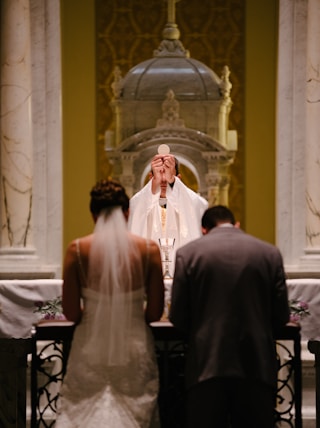 groom and bride kneeling in front of priest raising The Holy Sacrament