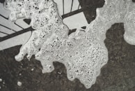 Bubbles and foam are spread unevenly over a dark surface, creating a textured pattern. The foam appears to be in various forms, with numerous small bubbles throughout, and is partially obscuring a geometric black and white design underneath.