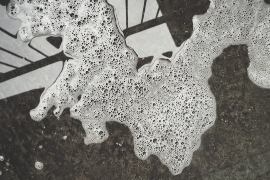 Bubbles and foam are spread unevenly over a dark surface, creating a textured pattern. The foam appears to be in various forms, with numerous small bubbles throughout, and is partially obscuring a geometric black and white design underneath.