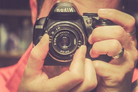 A close-up of hands holding a Nikon D3000 camera with the lens prominently in focus. The hands are adjusting the camera, and the background is softly blurred, emphasizing the foreground elements.