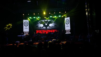 A dark stage with bright spotlights showcases a musical performance. The centerpiece is a backlit sign with a bat-like emblem and the word 'KERR' in red. Musicians are silhouetted against the light, contributing to a dramatic atmosphere.