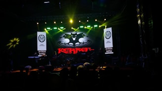 A dark stage with bright spotlights showcases a musical performance. The centerpiece is a backlit sign with a bat-like emblem and the word 'KERR' in red. Musicians are silhouetted against the light, contributing to a dramatic atmosphere.