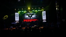 A dark stage with bright spotlights showcases a musical performance. The centerpiece is a backlit sign with a bat-like emblem and the word 'KERR' in red. Musicians are silhouetted against the light, contributing to a dramatic atmosphere.