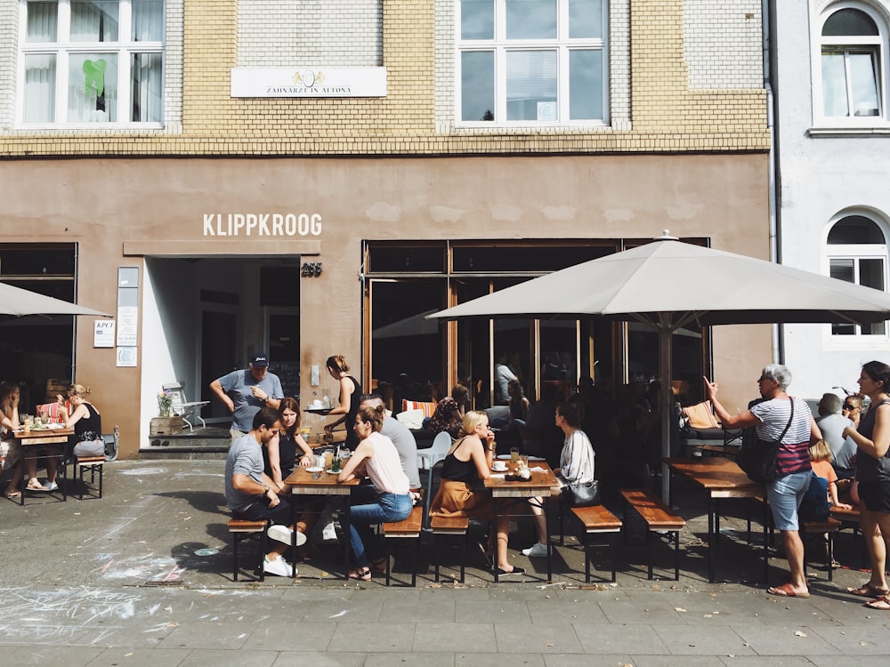 grupo de pessoas sentadas em bancos com mesas ao lado da frente da loja Klippkroog durante o dia