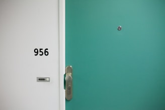 A green door with a silver door handle and a peephole. Adjacent to the door is a white wall with the number 956 and a small doorbell plate.