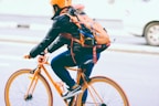 A person is riding a bicycle along a city street, wearing a helmet for safety. The bicycle and the helmet are both bright orange, matching the person's backpack. Dressed in casual attire, including a jacket and jeans, the cyclist appears focused and stable while holding the handlebars.