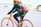 A person is riding a bicycle along a city street, wearing a helmet for safety. The bicycle and the helmet are both bright orange, matching the person's backpack. Dressed in casual attire, including a jacket and jeans, the cyclist appears focused and stable while holding the handlebars.