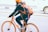 A person is riding a bicycle along a city street, wearing a helmet for safety. The bicycle and the helmet are both bright orange, matching the person's backpack. Dressed in casual attire, including a jacket and jeans, the cyclist appears focused and stable while holding the handlebars.