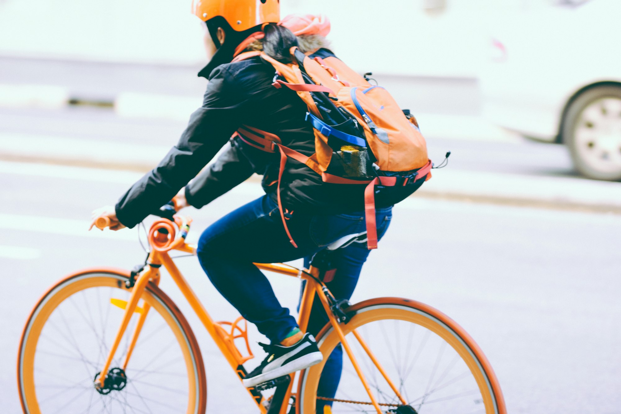 Une cycliste sur un vélo orange avec un sac à dos et un casque de la même couleur circulant en ville.