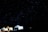 A starry night sky dominates the scene, with thousands of stars twinkling in the darkness. Below, several illuminated tents are pitched, creating a cozy and inviting atmosphere. The contrast between the bright tents and the dark sky highlights the tranquility of a night spent camping.