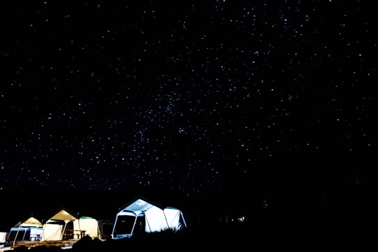 A starry night sky dominates the scene, with thousands of stars twinkling in the darkness. Below, several illuminated tents are pitched, creating a cozy and inviting atmosphere. The contrast between the bright tents and the dark sky highlights the tranquility of a night spent camping.