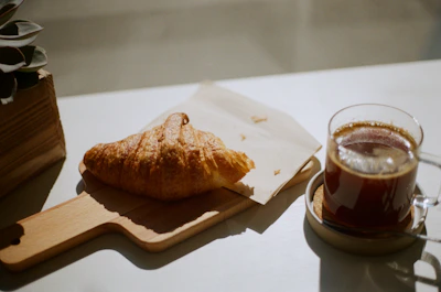 Warm morning light streaming through the coffee shop window onto a wooden table with a steaming cup of coffee and a croissant.