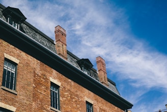 A brick building with multiple chimneys is set against a clear blue sky with wispy clouds. The architecture includes a slate roof and windows with dark frames.