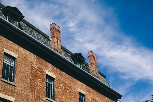 A brick building with multiple chimneys is set against a clear blue sky with wispy clouds. The architecture includes a slate roof and windows with dark frames.