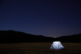 A bright tent is illuminated against the backdrop of a dark, starry night sky. Surrounding the tent is an expansive field and faint outlines of distant hills or mountains can be seen at the horizon.