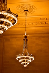A large, ornate chandelier with multiple tiers and intricate detailing is suspended from the ceiling. The ceiling features decorative moldings and a central medallion. The warm lighting creates an elegant and luxurious ambiance.