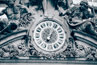An ornate clock with Roman numerals is surrounded by intricate sculptures and detailed carvings. The artwork includes figures and decorative elements, displaying high craftsmanship.