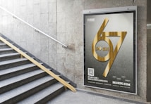 A staircase with a metal railing on the left leads upwards, surrounded by grey stone walls. On the right, there is a large advertisement poster featuring metallic golden numbers '67' with the years '1949-2016' embossed in the center. Below this, there is text along with a QR code and a website URL. The poster and its bold design stand out against the muted wall background.