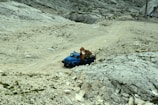 A blue utility truck equipped with a crane is parked on a rugged, rocky terrain that slopes upward. The area is sparsely vegetated, with some patches of small plants and grasses scattered across the rocky ground. In the background, there is a paved area with some equipment visible.