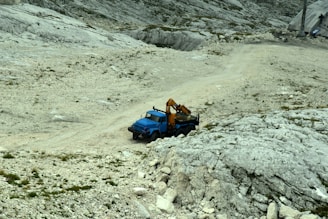 A blue utility truck equipped with a crane is parked on a rugged, rocky terrain that slopes upward. The area is sparsely vegetated, with some patches of small plants and grasses scattered across the rocky ground. In the background, there is a paved area with some equipment visible.
