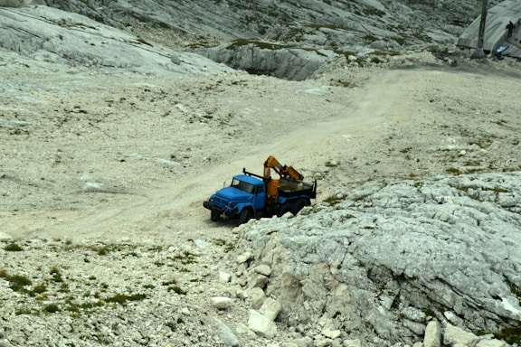 A blue utility truck equipped with a crane is parked on a rugged, rocky terrain that slopes upward. The area is sparsely vegetated, with some patches of small plants and grasses scattered across the rocky ground. In the background, there is a paved area with some equipment visible.