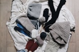 A variety of knitted and crocheted textiles, including hats, scarves, and socks, arranged on a wooden floor. There are several balls of yarn in different colors, knitting needles, and crochet hooks scattered around. The textures are soft and cozy, featuring earthy tones like gray, beige, and dark blue.