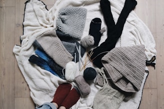 A variety of knitted and crocheted textiles, including hats, scarves, and socks, arranged on a wooden floor. There are several balls of yarn in different colors, knitting needles, and crochet hooks scattered around. The textures are soft and cozy, featuring earthy tones like gray, beige, and dark blue.