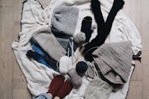 A variety of knitted and crocheted textiles, including hats, scarves, and socks, arranged on a wooden floor. There are several balls of yarn in different colors, knitting needles, and crochet hooks scattered around. The textures are soft and cozy, featuring earthy tones like gray, beige, and dark blue.