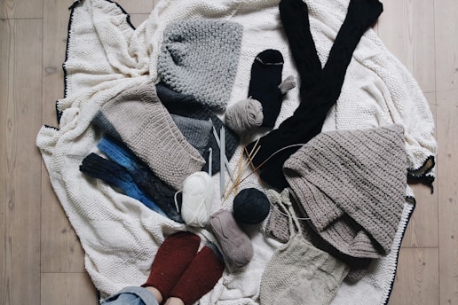 A variety of knitted and crocheted textiles, including hats, scarves, and socks, arranged on a wooden floor. There are several balls of yarn in different colors, knitting needles, and crochet hooks scattered around. The textures are soft and cozy, featuring earthy tones like gray, beige, and dark blue.