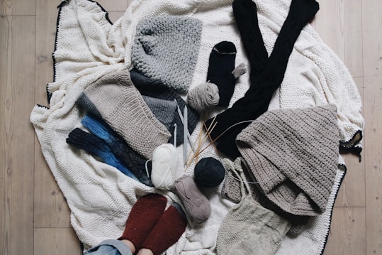 A variety of knitted and crocheted textiles, including hats, scarves, and socks, arranged on a wooden floor. There are several balls of yarn in different colors, knitting needles, and crochet hooks scattered around. The textures are soft and cozy, featuring earthy tones like gray, beige, and dark blue.