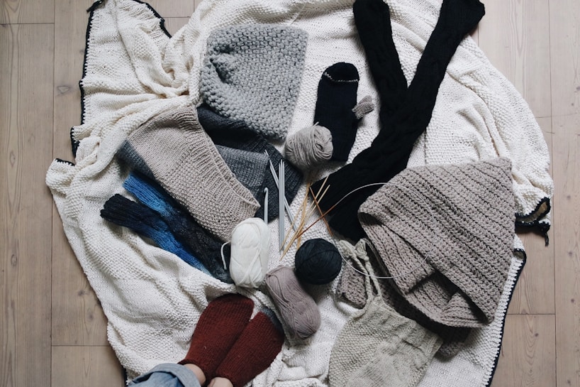 A variety of knitted and crocheted textiles, including hats, scarves, and socks, arranged on a wooden floor. There are several balls of yarn in different colors, knitting needles, and crochet hooks scattered around. The textures are soft and cozy, featuring earthy tones like gray, beige, and dark blue.